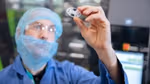 A gloved hand holding a small, circular plastic component in a cleanroom environment with machinery in the background.