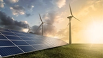 Solar panels installed on a grassy field with wind turbines in the background under a partly cloudy sky and bright sunlight.