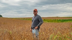 A farmer stands in a field of crops, looking at the camera. He is wearing blue jeans and a red baseball hat.