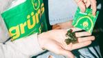 Green gummy supplements resting in an open hand, with more gummies being poured from a small Grüns packet; a larger Grüns pouch is visible in the background.